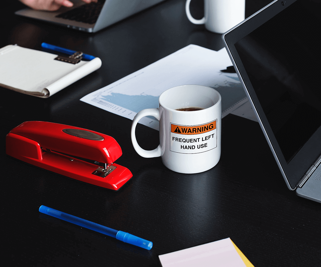 White ceramic mug for left-handers with a warning frequent left hand use hazard sign design on a desk during business meeting.